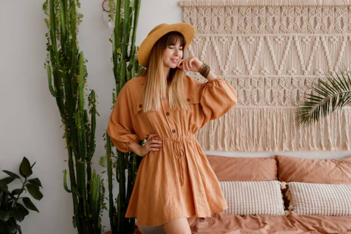 A person poses in a stylish, buttoned, peach-colored dress with a cinched waist, surrounded by green plants and a cozy bohemian setting.