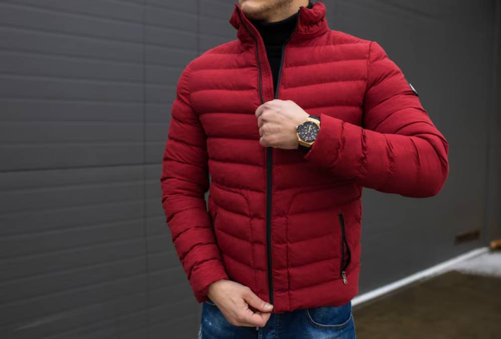 A man in a red puffer jacket zips up his coat, showcasing a black turtleneck and a stylish watch against a neutral gray background.