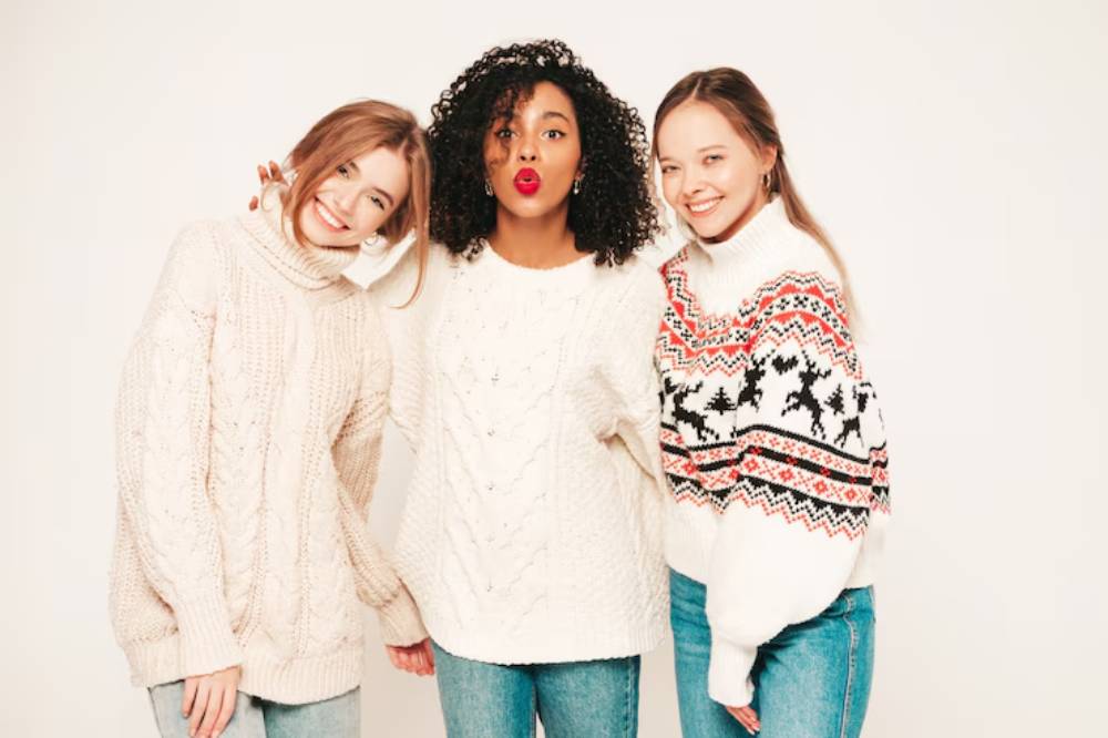 Three women, each wearing cozy knit sweaters in neutral tones and festive patterns, stand together against a plain white background.
