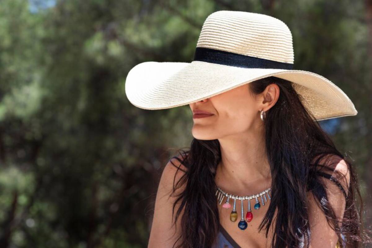 A woman with long dark hair wearing a wide-brimmed straw hat and a colorful necklace sits outdoors against a blurred green backdrop.