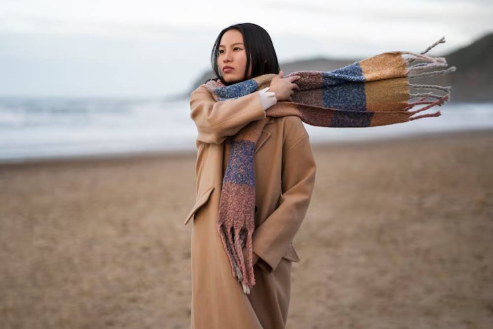 A person in a long beige coat stands on a beach, playfully draping a colorful scarf as ocean waves crash in the background.