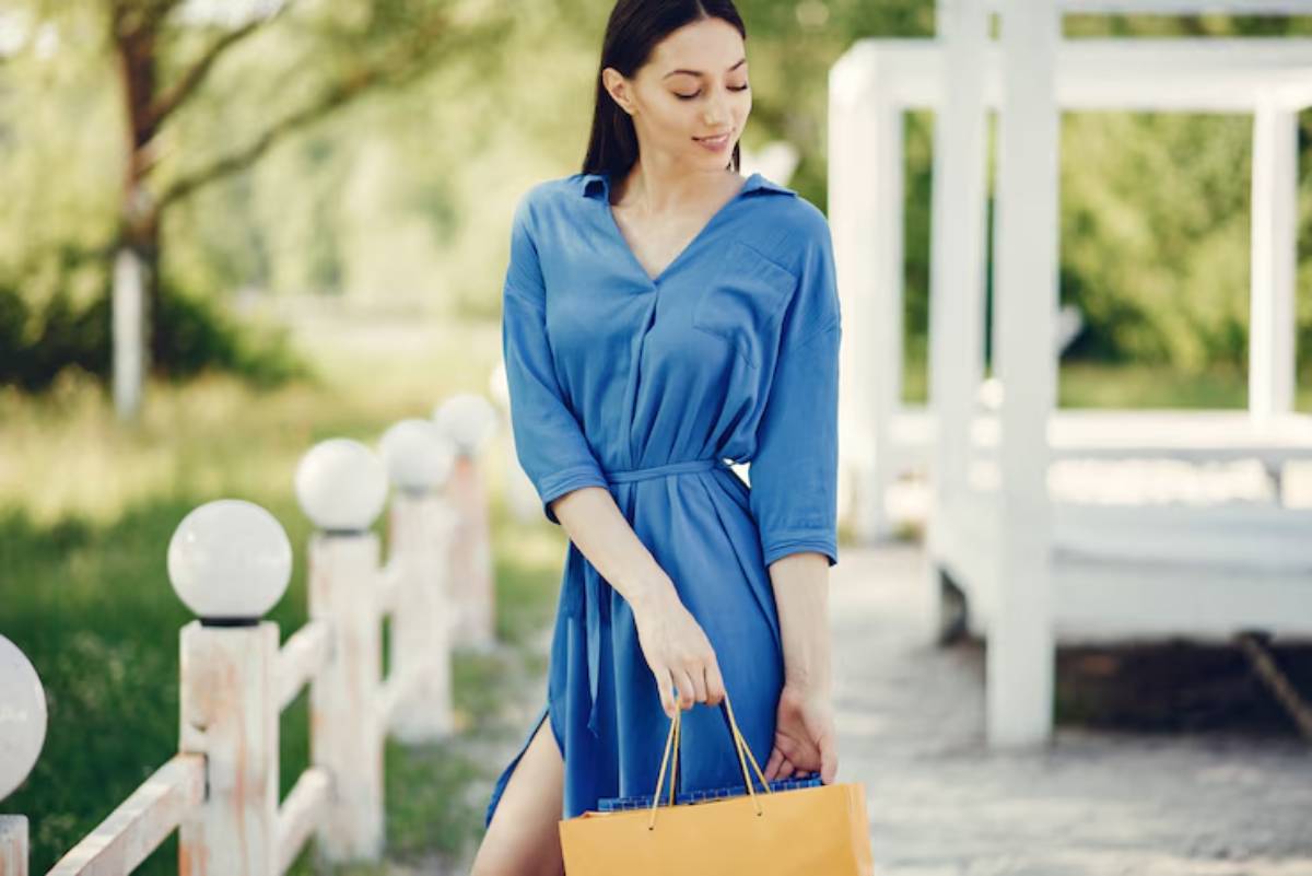 A woman in a blue shirt dress holds a yellow handbag while standing on a path in a greenery-filled outdoor setting.