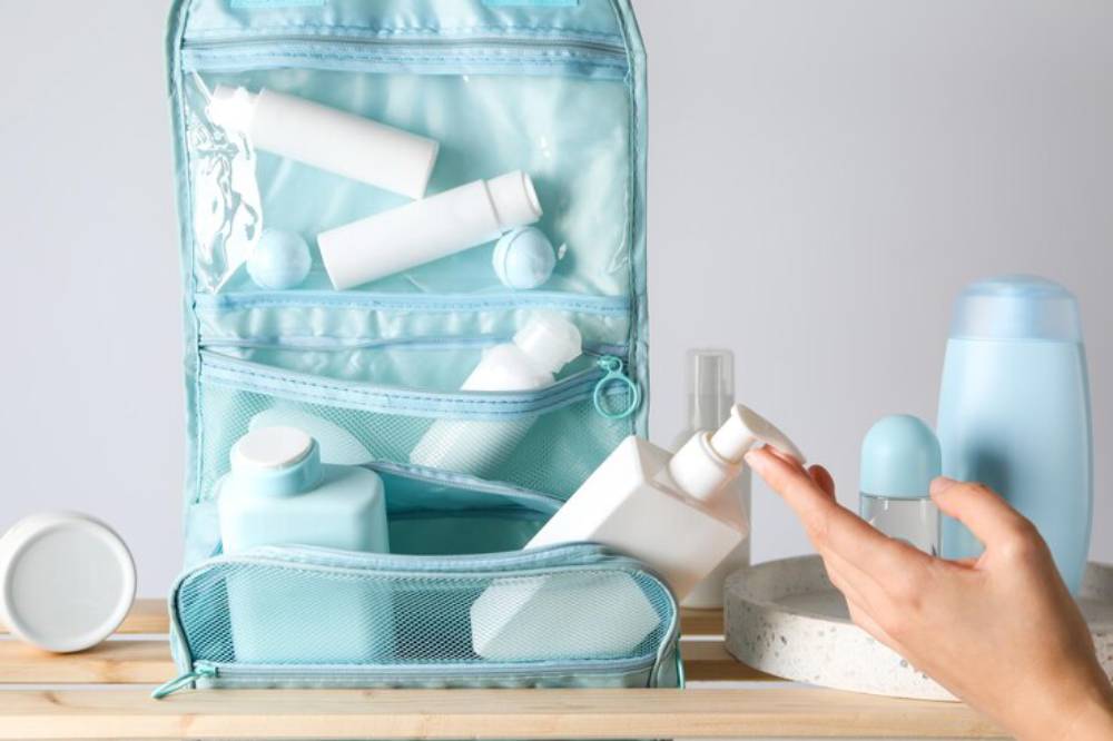 A hand reaching for a white bottle from a light blue travel toiletries bag filled with various containers and bottles.