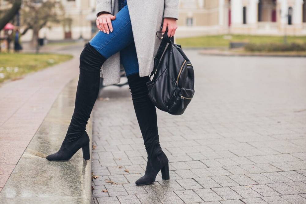 A woman in high black boots and blue jeans stands on a sidewalk, holding a black backpack, with a city background.