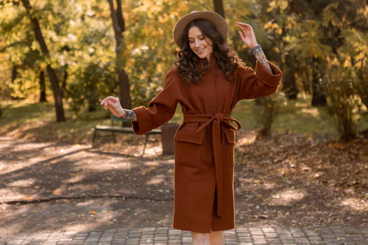 A woman in a Spiced-Caramel colored coat and hat strikes a playful pose amidst a sunlit, autumnal park setting.
