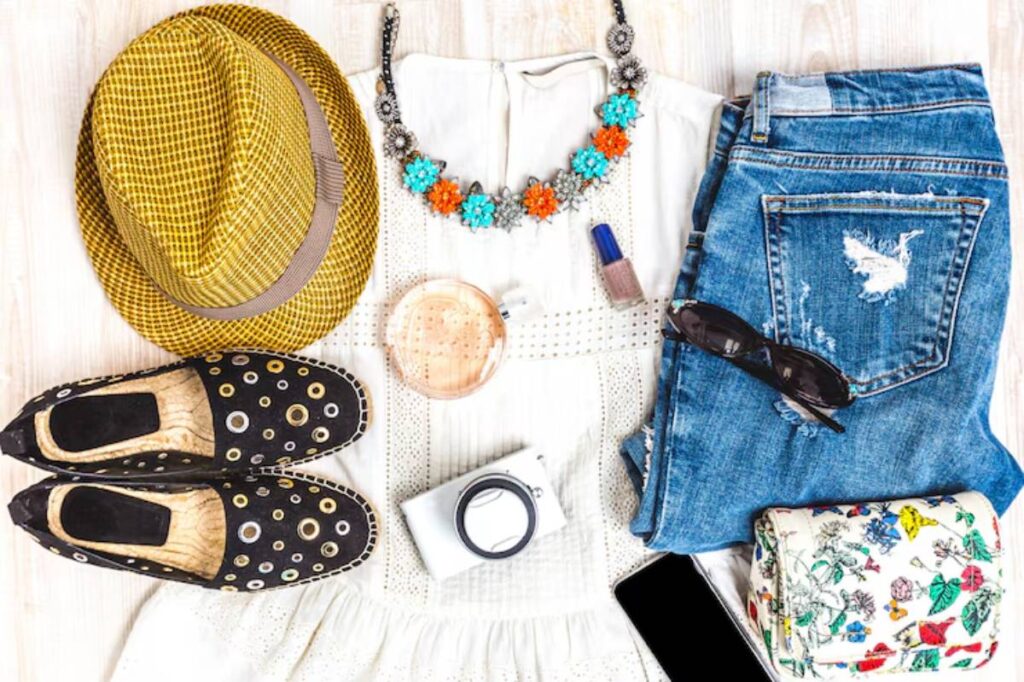 A stylish flat lay featuring a yellow hat, floral necklace, denim, black shoes, camera, sunglasses, and a floral wallet.