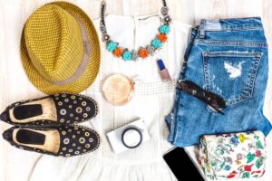 A stylish flat lay featuring a yellow hat, floral necklace, denim, black shoes, camera, sunglasses, and a floral wallet.