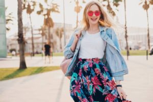 A woman in a floral skirt and denim jacket walks through a sunny park lined with palm trees, exuding a carefree vibe.