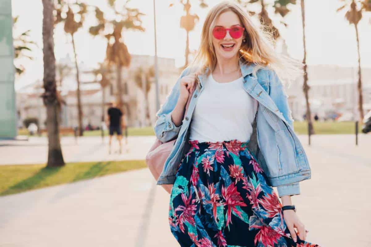 A woman in a floral skirt and denim jacket walks through a sunny park lined with palm trees, exuding a carefree vibe.