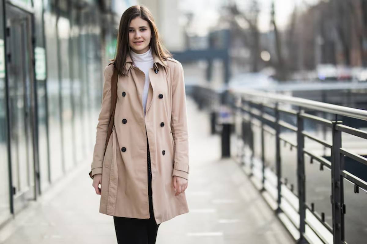 A woman in a stylish beige trench coat walks along a sunlit urban walkway, with modern buildings in the background.