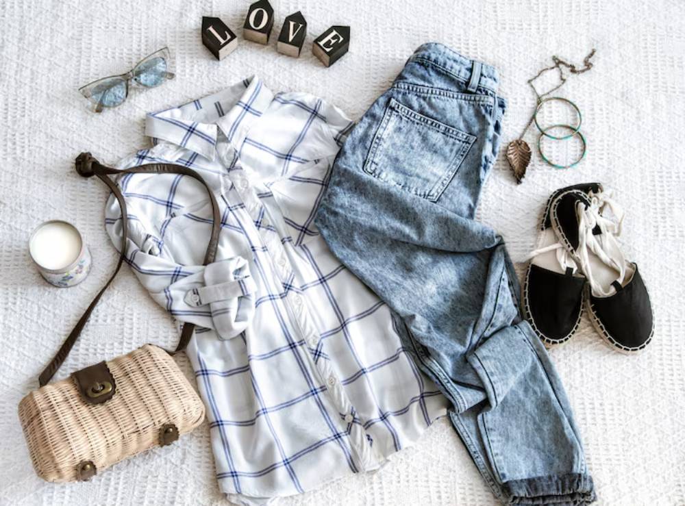 Flat lay of a casual outfit: plaid shirt, denim jeans, black espadrilles, sunglasses, a bag, and accessories on a textured white background.
