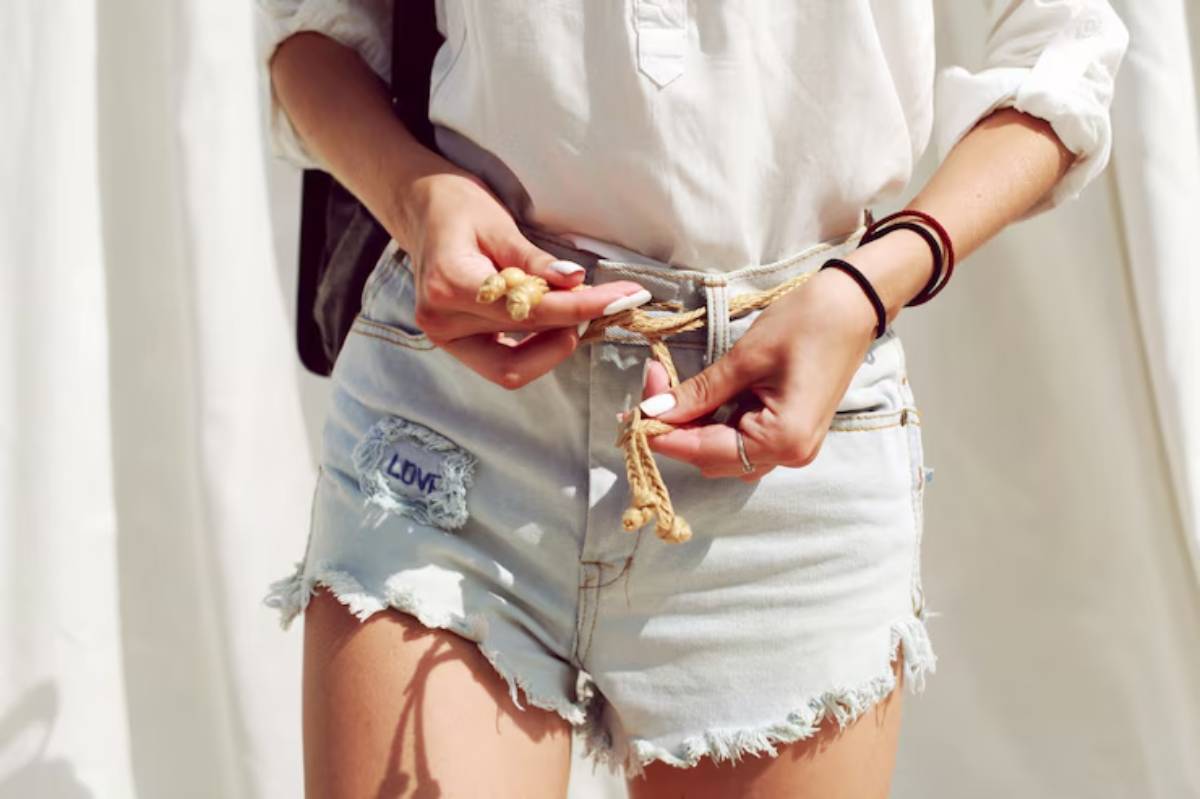 A woman in a white blouse adjusts the rope belt of her distressed denim shorts, showcasing a casual summer style.