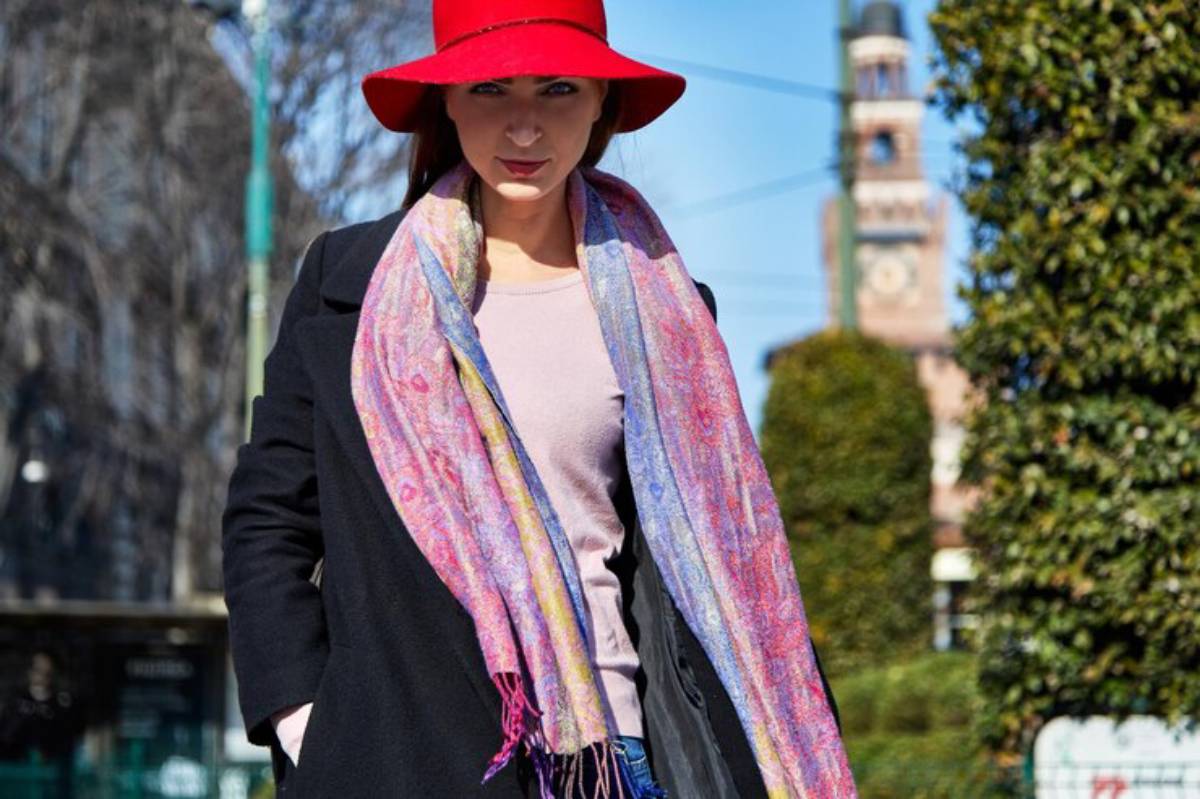 A woman in a black coat and a vibrant pink scarf stands outdoors, with trees and an architectural tower visible in the background.