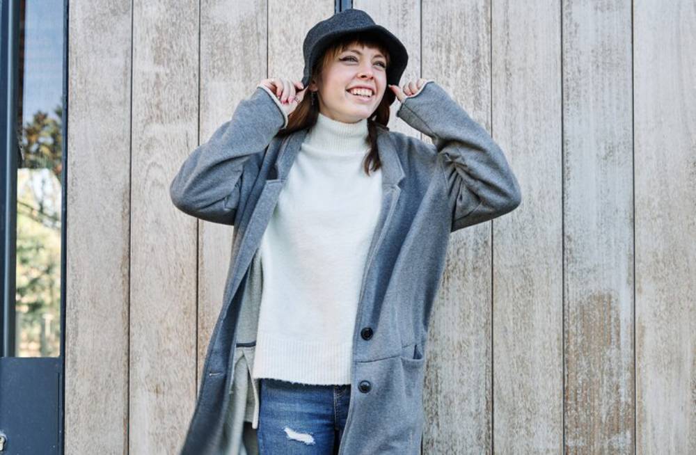 A person poses outdoors, wearing a gray long coat and a hat, in front of a wooden wall. They are styled casually with ripped jeans.