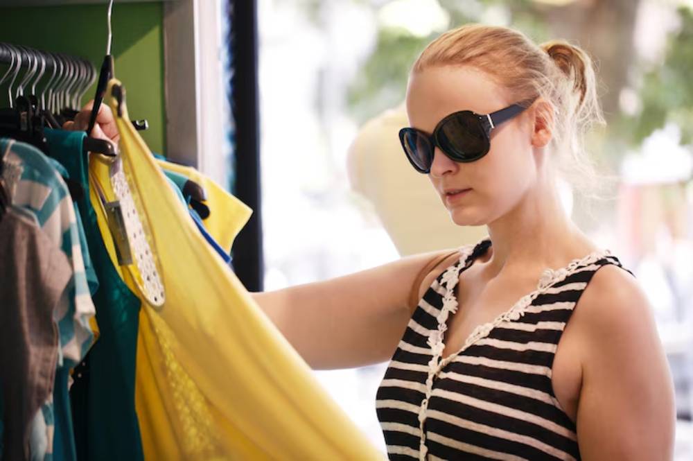 A woman browses colorful clothing on a rack in a bright, inviting store, holding a yellow dress while wearing stylish sunglasses.