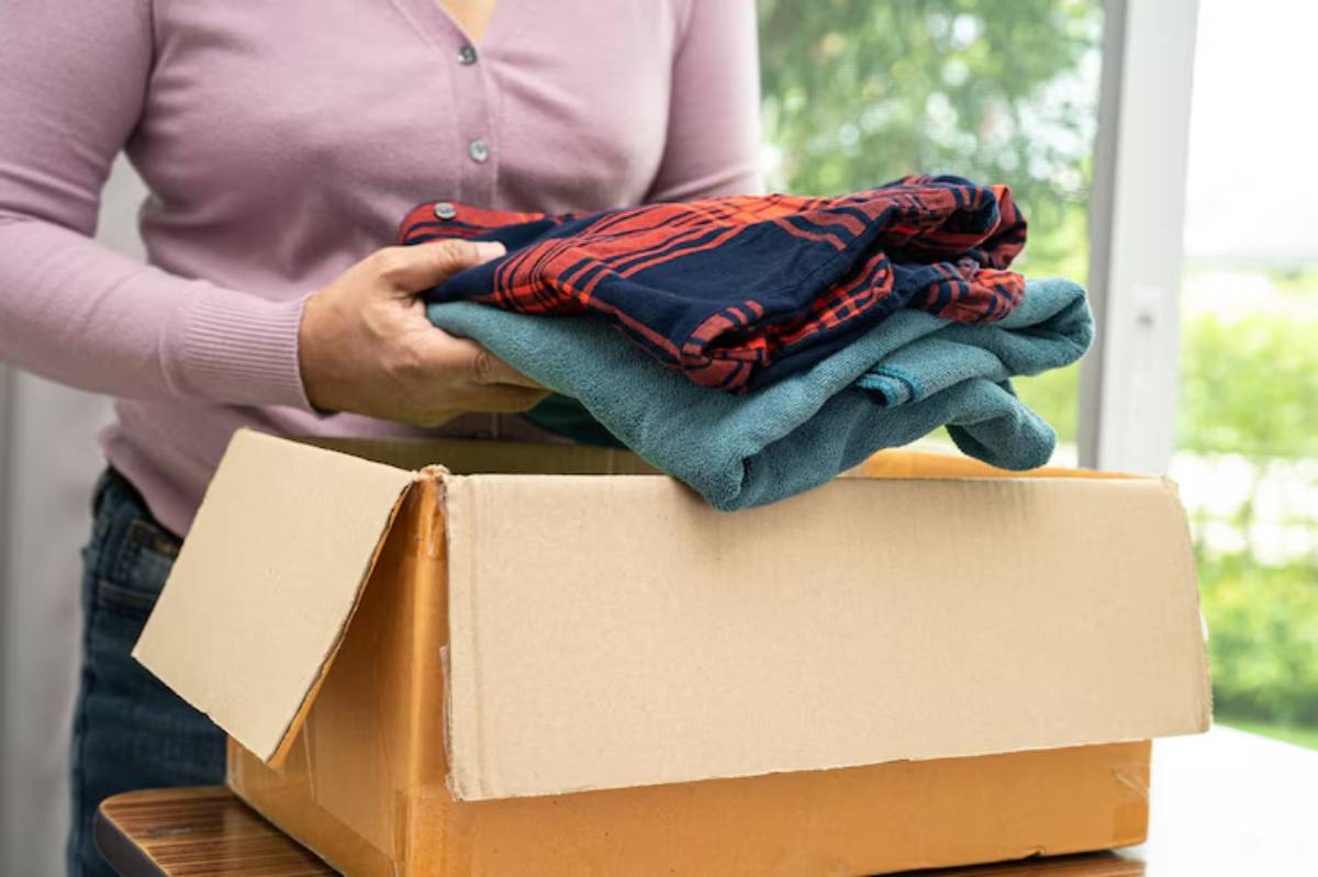 A person in a purple cardigan places folded clothes into a cardboard box, preparing for packing.