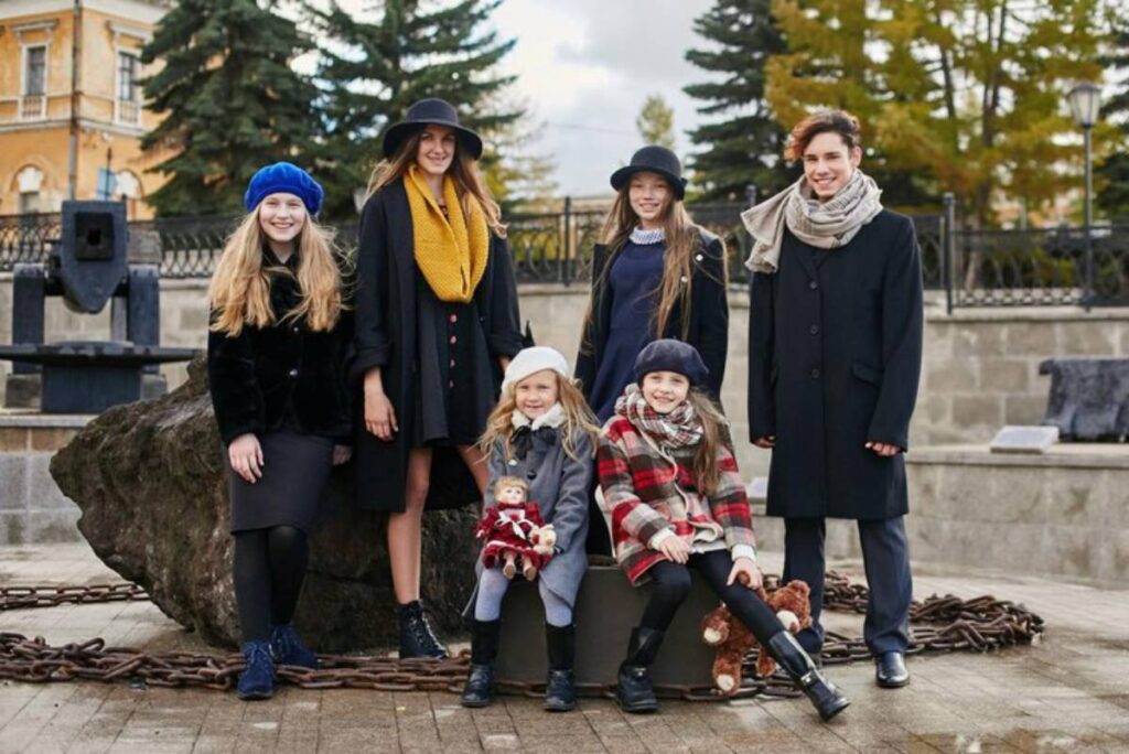 A group of stylishly dressed individuals poses outdoors near a large rock and chains, with trees and historical architecture in the background.