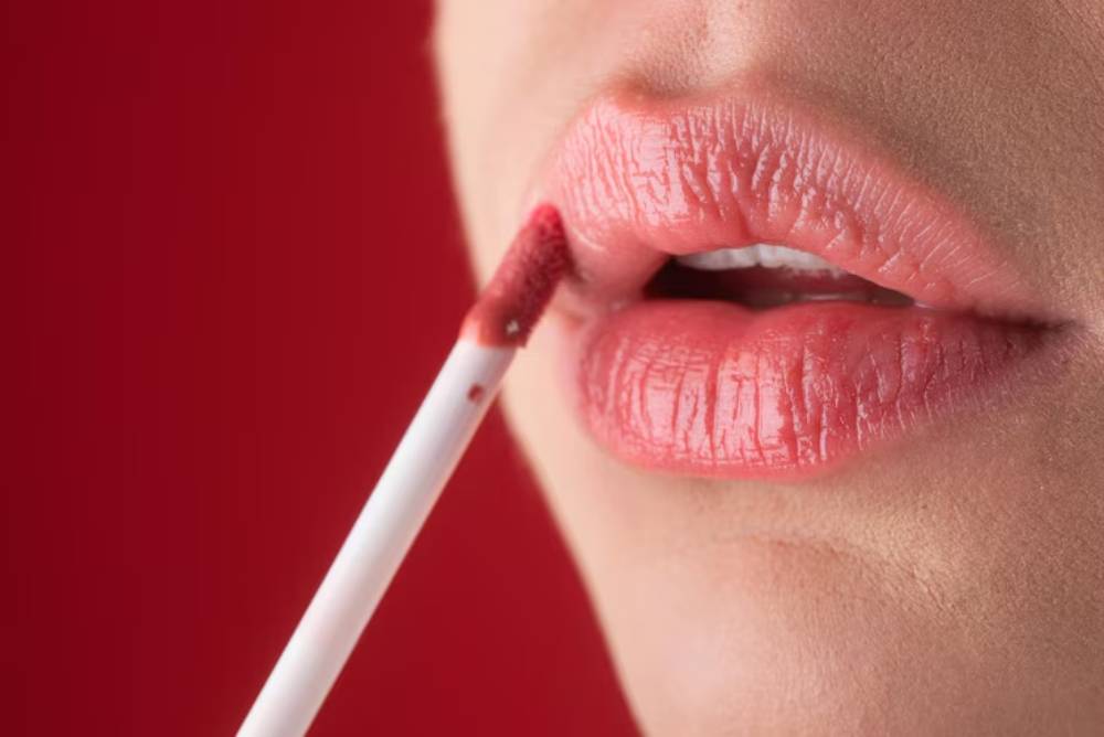 Close-up of lips applying a soft pink lip gloss with a wand, set against a vibrant red background.