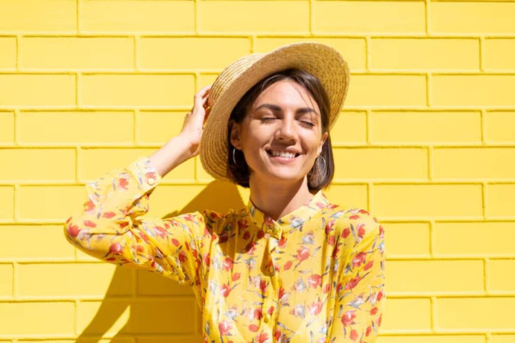 A person wearing a bright floral yellow blouse and a straw hat stands against a vibrant yellow brick wall, exuding a cheerful vibe.