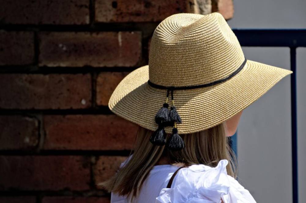 A woman with long hair wears a wide-brimmed straw hat adorned with black tassels, standing against a brick wall background.