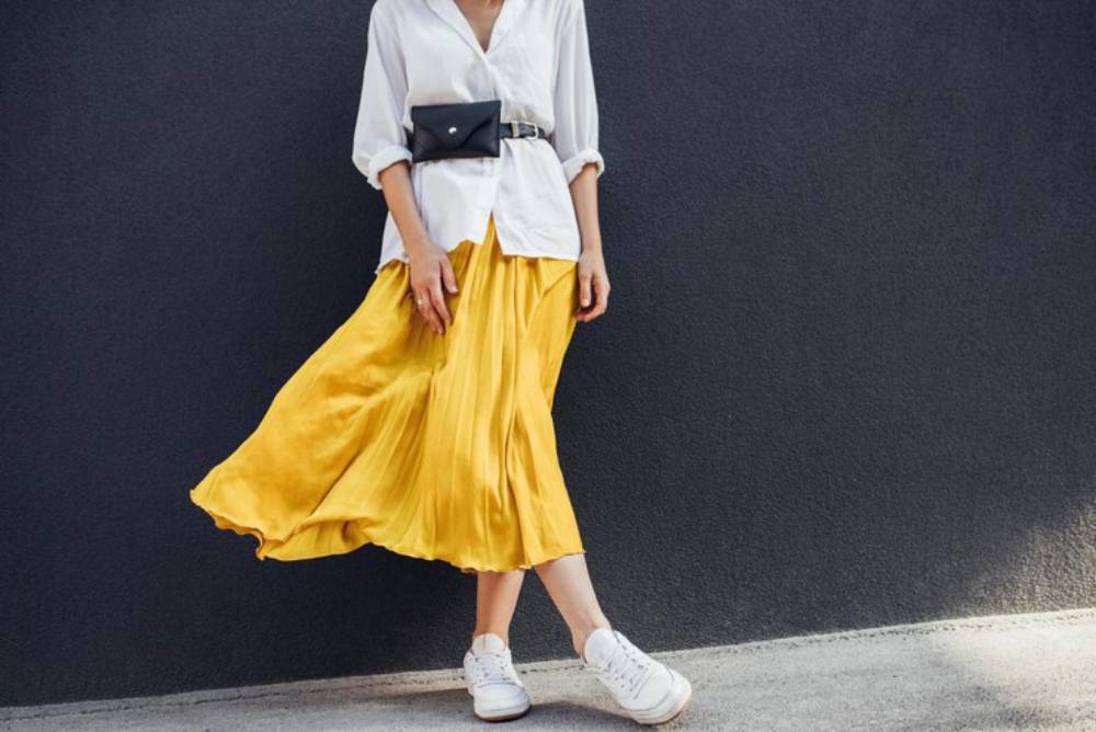 A woman in a white shirt and yellow pleated skirt stands against a dark wall, completing her look with white sneakers and a belt bag.