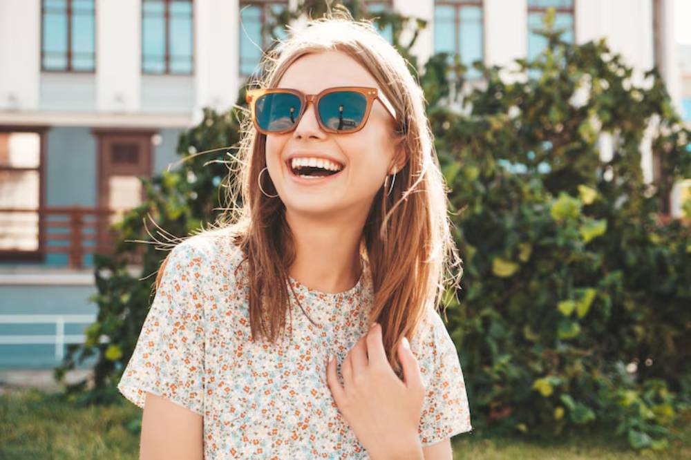 A young person with long hair is wearing a floral shirt and a sunglasses, standing outdoors in front of greenery and a light-colored building.