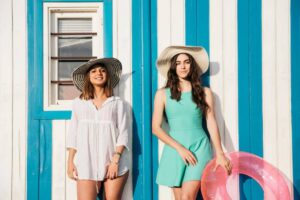 Two women stand in front of a blue and white striped wall, wearing swimsuits and large sun hats. One holds a pink inflatable ring.