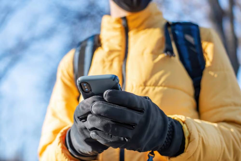 A person in a yellow jacket and gloves holds a smartphone while outdoors on a sunny day, with trees in the background.