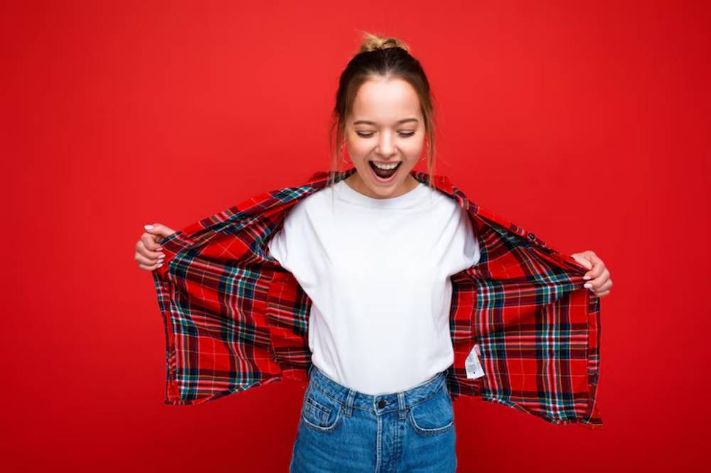 A person wearing a white t-shirt and denim jeans, holding open a red plaid shirt against a bright red background.