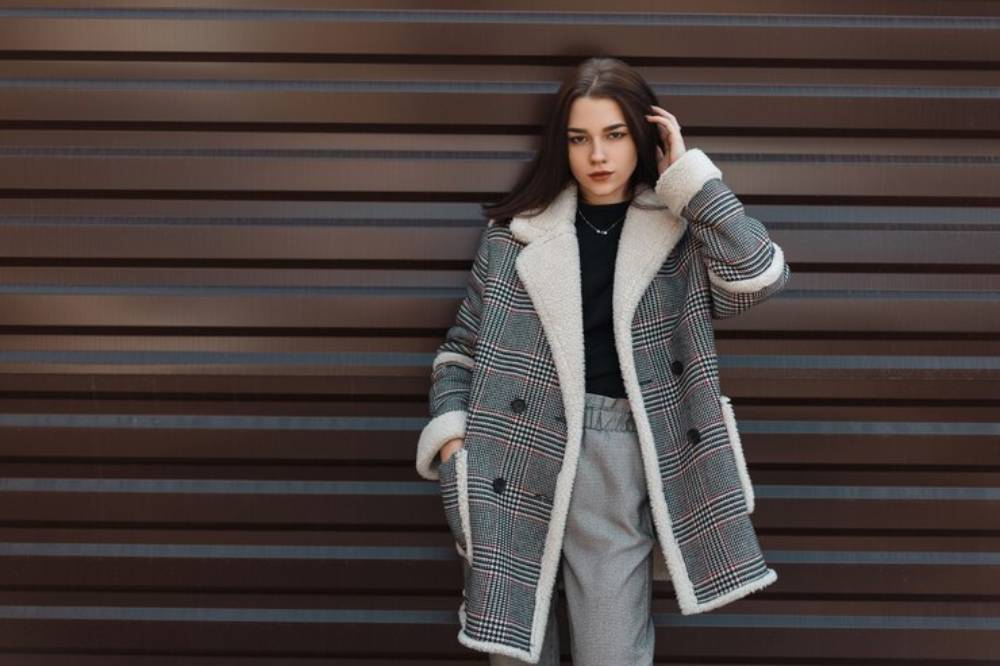A person stands against a striped brown wall, wearing a stylish plaid coat with a cozy lining and light-colored trousers.