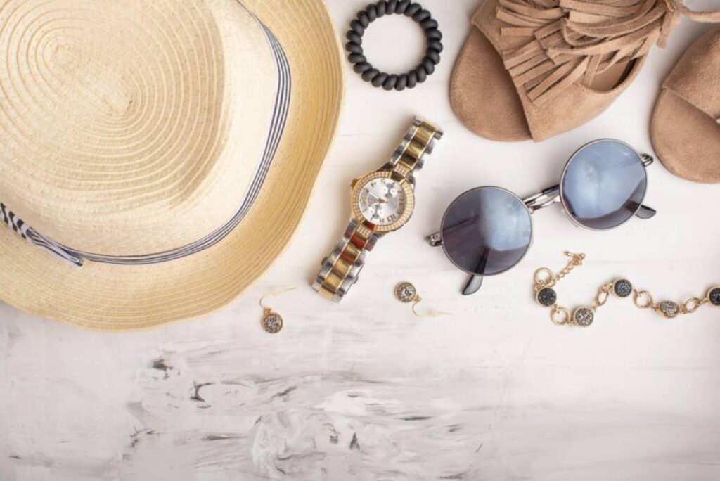Flat lay of summer accessories: a straw hat, sunglasses, a stylish watch, earrings, a bracelet, and beige sandals on a white surface.
