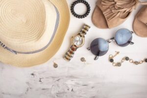 Flat lay of summer accessories: a straw hat, sunglasses, a stylish watch, earrings, a bracelet, and beige sandals on a white surface.