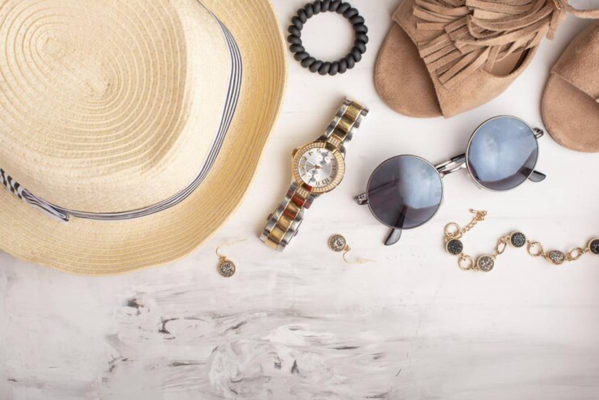 Flat lay of summer accessories: a straw hat, sunglasses, a stylish watch, earrings, a bracelet, and beige sandals on a white surface.