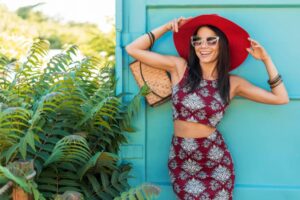 A woman in a burgundy floral two-piece outfit poses against a turquoise wall, accessorized with a wide-brimmed red hat and a bag.