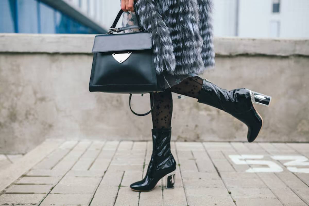 A stylish individual walks on pavement, dressed in grey fur, black patent boots, and lace star-patterned tights, holding a chic black handbag.