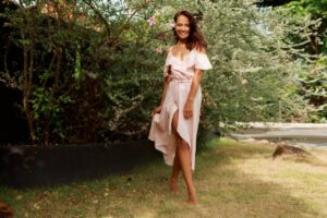 A person stands gracefully in a light pink dress with a high slit, surrounded by lush greenery and soft sunlight.
