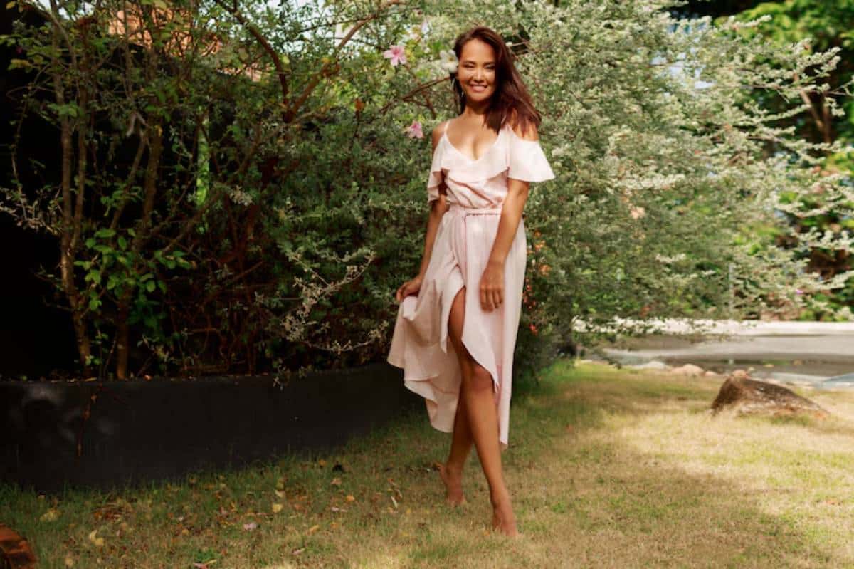 A person stands gracefully in a light pink dress with a high slit, surrounded by lush greenery and soft sunlight.