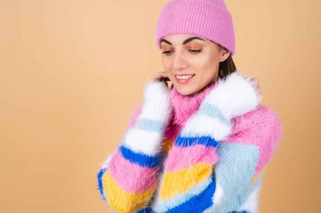 A person in a vibrant, multicolored striped sweater and a pink beanie stands against a soft orange background, looking cozy and stylish.