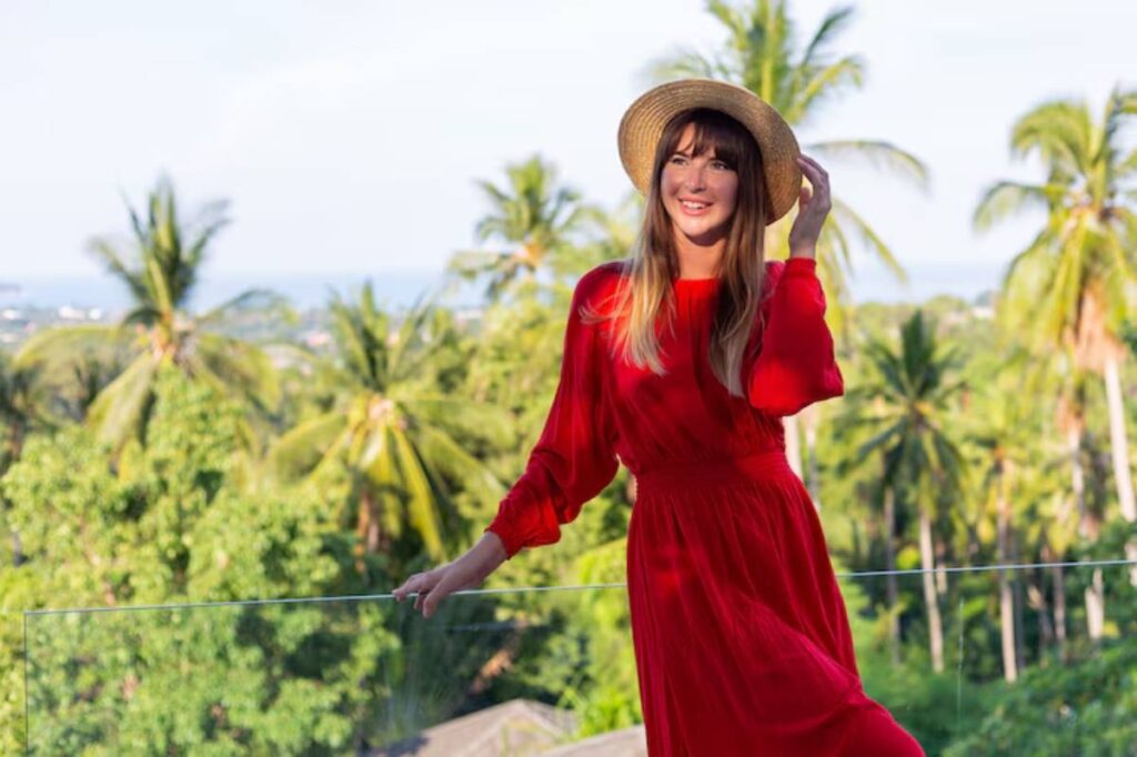 A woman in a flowing red dress and straw hat poses against a lush tropical backdrop of palm trees and a distant ocean view.