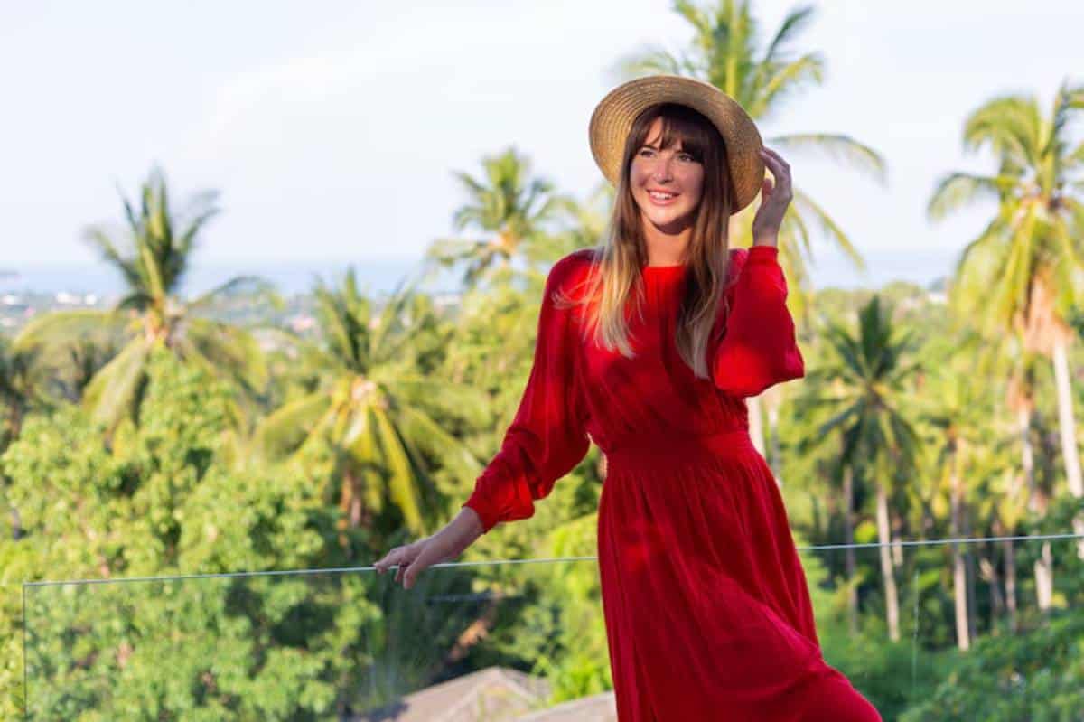 A woman in a flowing red dress and straw hat poses against a lush tropical backdrop of palm trees and a distant ocean view.